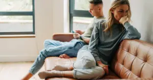 A woman looking away in deep thought, while a man behind her focuses on his phone, symbolizing emotional disconnection and avoidance in relationships.