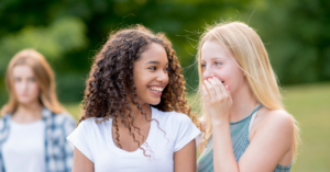 Two preteen girls gossiping about another girl sitting alone, highlighting the social challenges of neurodiversity and ableism.