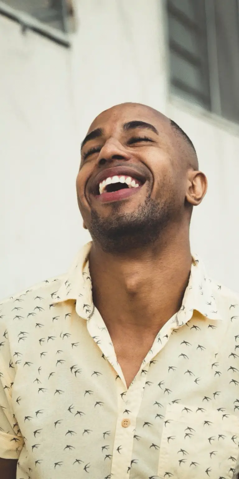 Close-up of a joyful man laughing with his head tilted back, wearing a light shirt with a bird pattern. A moment of lightness, relief, and release.